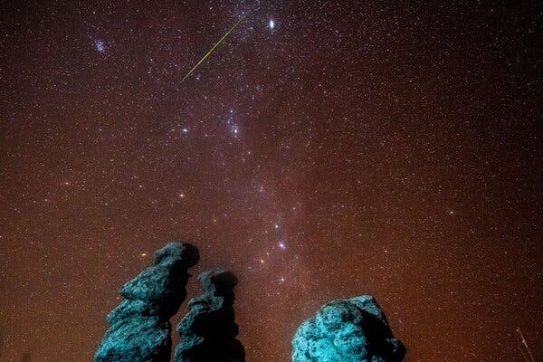 A streak of light flies through a starry sky over blue-green rock formations.