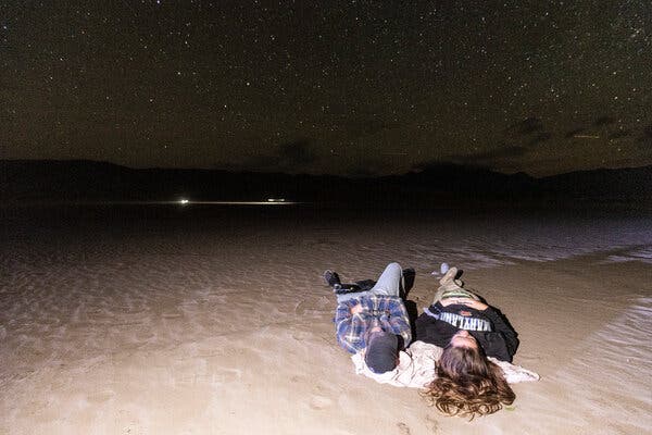 Two people lying on a beach, staring up at a night sky full of stars.