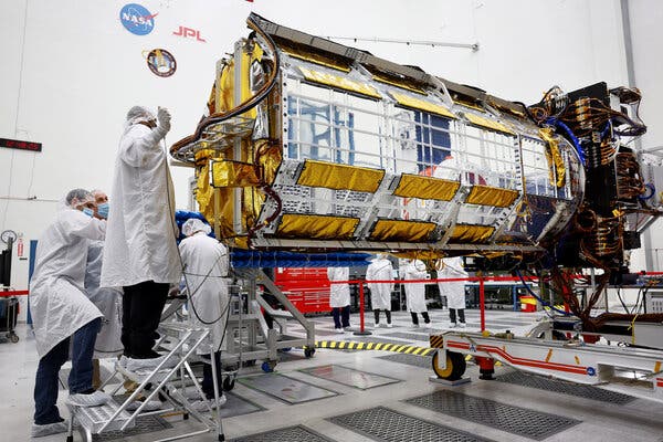 Several scientists or technicians in white lab coats and masks and hairnets tend to a large metallic spacecraft in a clean room.