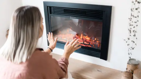 Getty Images Woman with blonde hair in pink-ish top warms her hands on a gas fire 