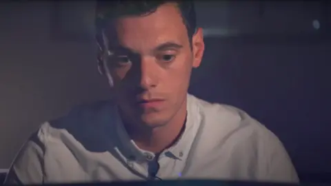 BBC A man with brown hair and wearing a light blue shirt looks in contemplation.