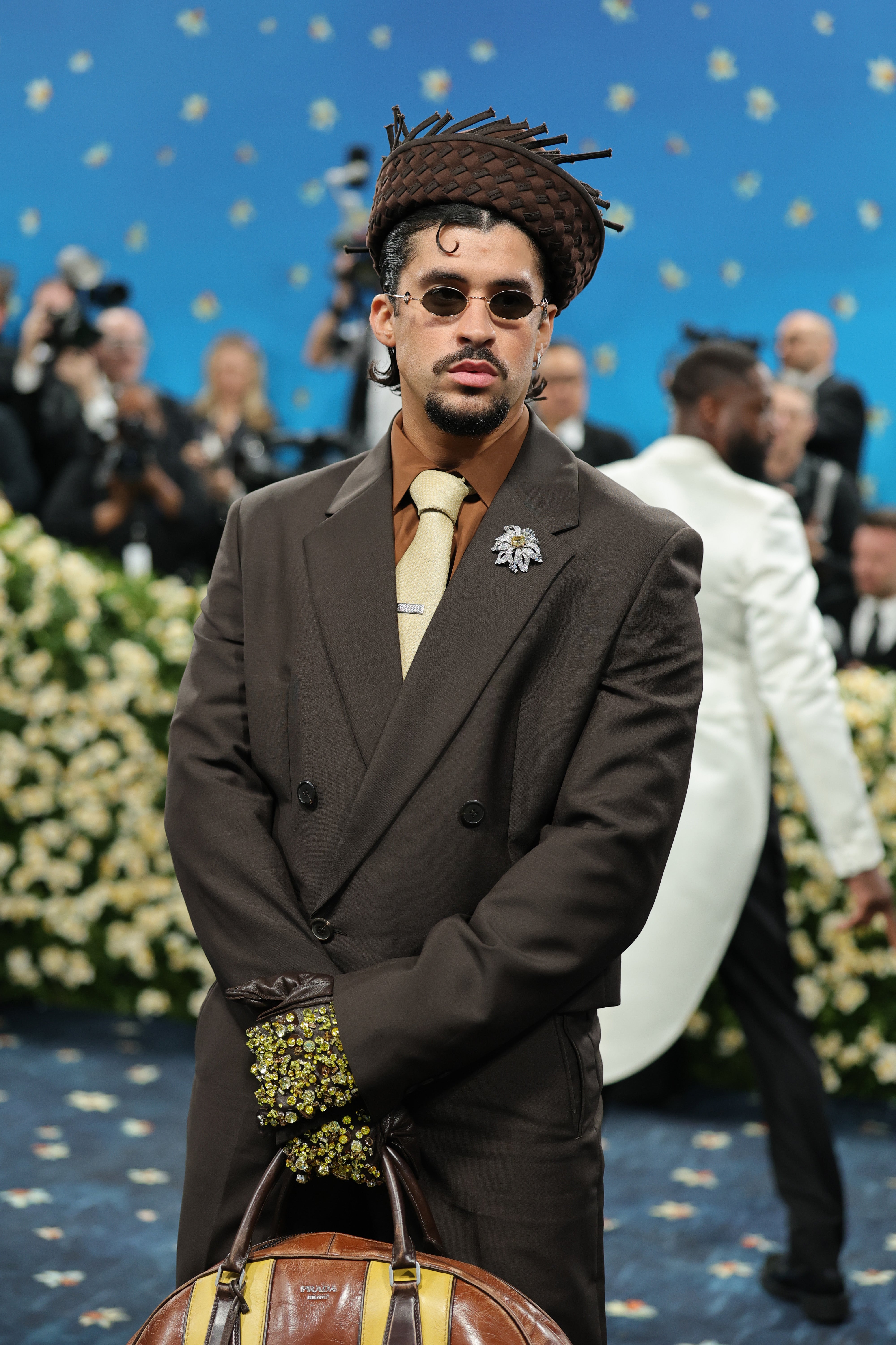 Bad Bunny stunned in a brown suit and beige tie by Prada. He paired the jacket, which featured a white brooch, with a brown straw hat