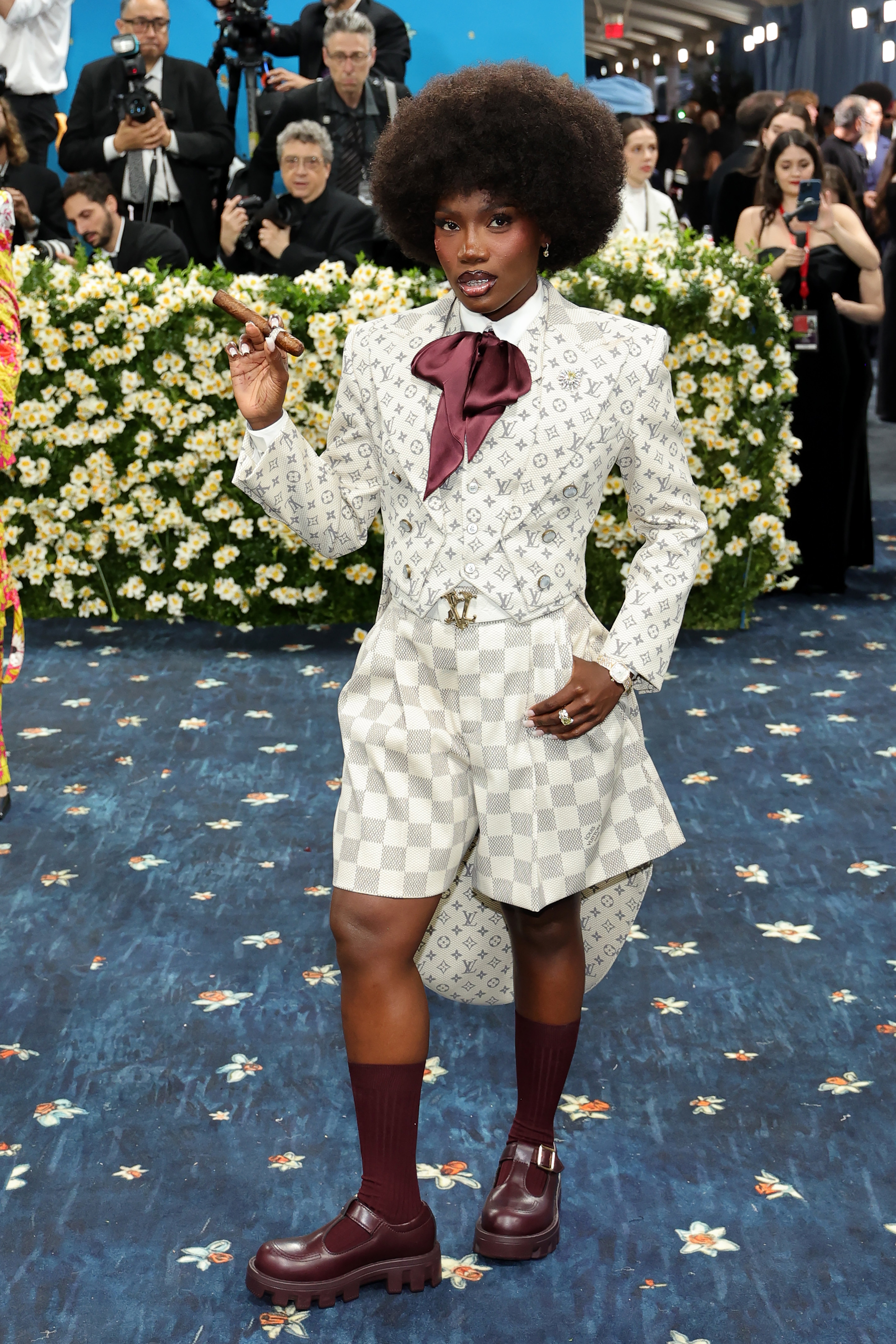 Doechii wore a white Louis Vuitton jacket and checkerboard shorts. She had a satin purple bowtie attached to her shirt and a cigar in her hand. She completed the look with maroon sandals and matching knee-high socks