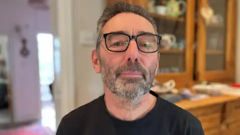 BBC Wearing glasses and a black T-shirt, Peter Holden poses for a photograph in his kitchen at his Worcestershire home. He has the start of a beard with dark and white hair.