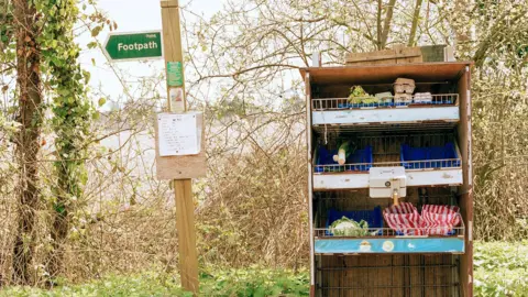 Kathryn Martin A small, shelved honesty box containing eggs, leeks and a cauliflower, and with a cash box fixed to the front, stands next to a footpath sign in what appears to be a rural setting.