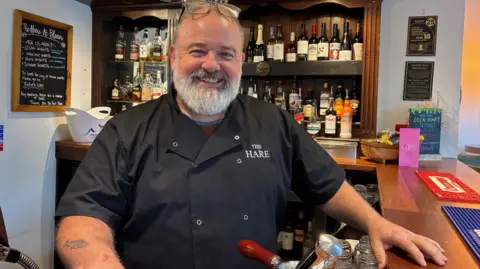 Matthew Hayden stands behind the bar of his pub wearing a chef's top branded with The Hare pub name.