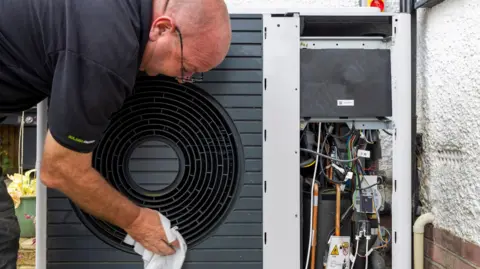 Andrew Aitchison/Getty Images An engineer - a man wearing a black T-shirt - wipes the outside of a black heat pump with a cloth. To the right of the picture the heat pump abuts the wall of a property with the pipes and electrical components visible