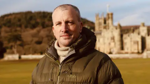 Dr Langlands wears a brown coat and stands in a field with a mountain and building in the background, he looks at the camera with a closed-mouth smile. It is a head and shoulders shot.