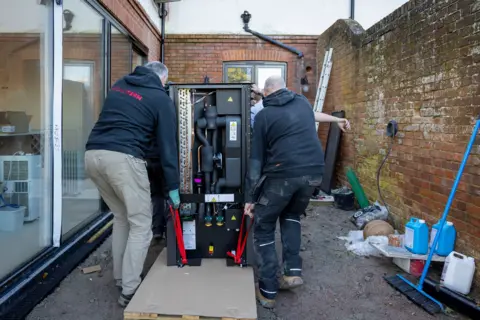 Andrew Aitchison/Getty Images Three men with their backs to the camera are shown lifting a black heat pump on a trolley towards the back of the house. The men wear dark work clothes and boots. The house is red brick with a long glass door to the left and the property boundary wall to the right. Against the wall sits work tools and a blue broom. 